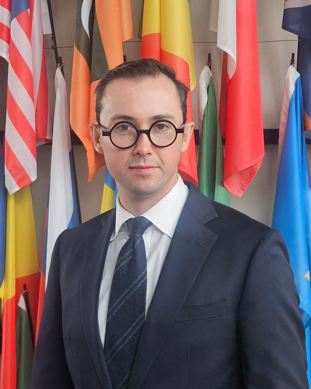 Fennar Ralston in a dark suit and round glasses, standing in front of a wall of national flags.